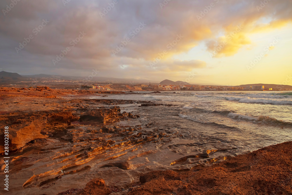 Golden overcast sunrise over El Medano town, in Tenerife with the limestone coast in the foreground. Mellow light sunrise over the Atlantic with calm waters, rocky shores and the town in the distance.