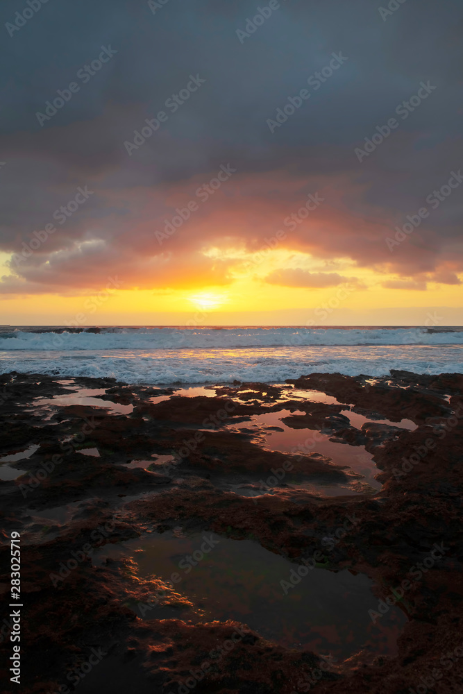 Vertical perspective of tropical sunrise over the limestone coasts of El Medano, Tenerife, Canary Islands, Spain. Seascape with vibrant colors, golden haze and dynamic waves striking rocky shores. Vertical perspective of tropical sunrise over the limestone coasts of El Medano, Tenerife, Canary Islands, Spain. Seascape with vibrant colors, golden haze and dynamic waves striking rocky shores.