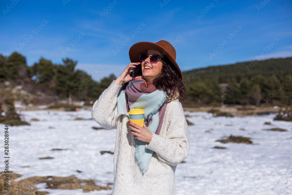 young beautiful woman talking on her mobile phone in the snow and holding a cup of tea or coffee. Outdoors portrait, lifestyle