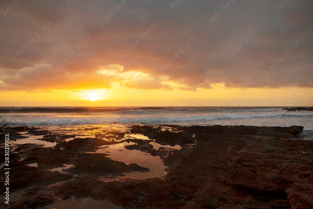 Glorious sunrise reflected in the natural water pools formed in the limestone shores of El Medano resort, Tenerife, Canary Islands, Spain, vibrant seascape with overcast sky and golden haze light