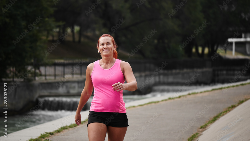 Beautiful adult red haired woman athlete in pink top and shorts makes amorning run on the city bridge over the river. Healthy lifestyle, jogging
