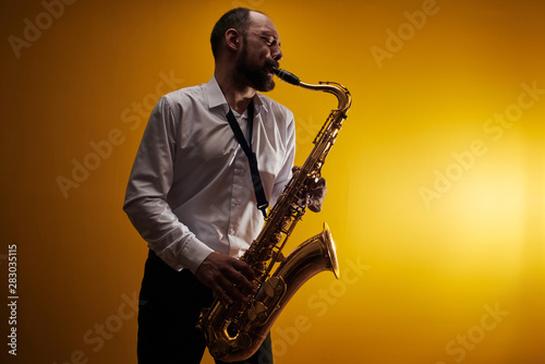 Portrait of professional musician saxophonist man in  white shirt plays jazz music on saxophone  yellow background in a photo studio  side view