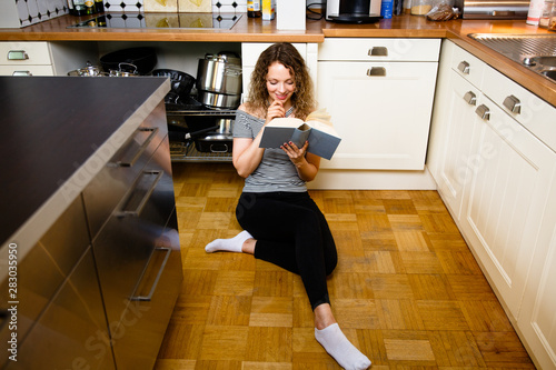 Hübsche junge Frau liest ein Buch in der Küche, sitzend auf dem Fußboden.