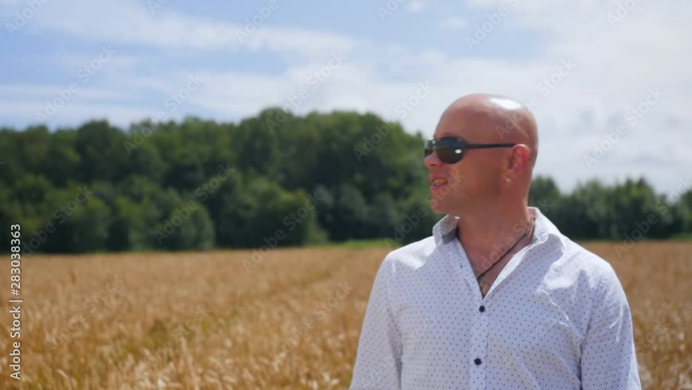 Portrait shot of a bald man in the white shirt standing in the golden wheat field, singing on the sunny sky background. Medium shot. Portrait shot of a bald man in the white shirt standing in the golden wheat field, singing on the sunny sky background. Medium shot.