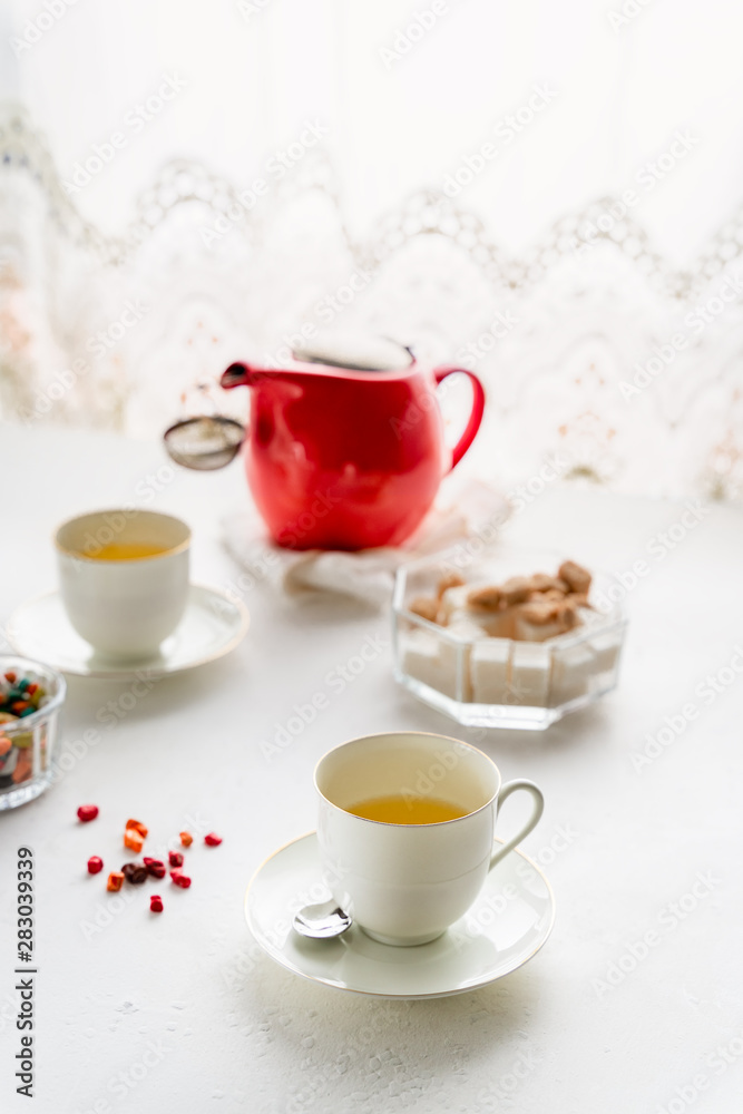 Red tea pot, two white tea cups, sugar bowl on a table by window Red tea pot, two white tea cups, sugar bowl on a table by window
