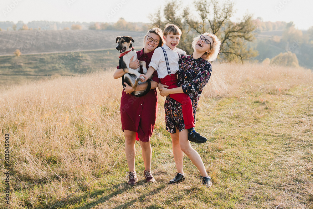Fototapeta premium Samesex caucasian lesbian family with a child and a dog walking outdoors on the background of beautiful nature. Mothers having fun with their son.