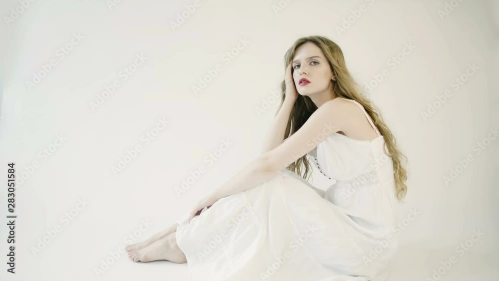 Girl with long hair in white long dress sits on white background in studio. Delicate facial features, nude makeup. Wind blows hair. She touches her hair, straightens her hair. Dreaming. Girl with long hair in white long dress sits on white background in studio. Delicate facial features, nude makeup. Wind blows hair. She touches her hair, straightens her hair. Dreaming.