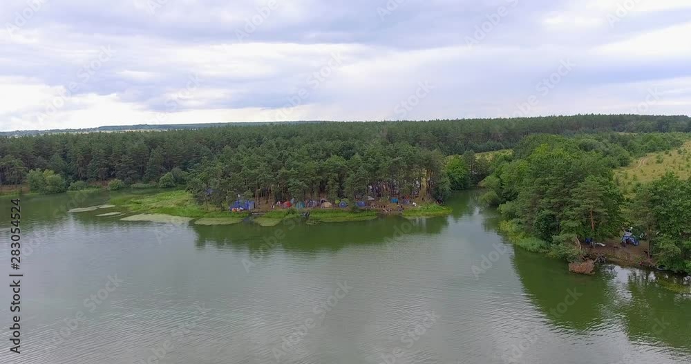 Aerial view. Camping at summers festival near the river in the pine forest. Festival or camp where many colorful tents. Flying low above the river and beautiful green forest Aerial view. Camping at summers festival near the river in the pine forest. Festival or camp where many colorful tents. Flying low above the river and beautiful green forest