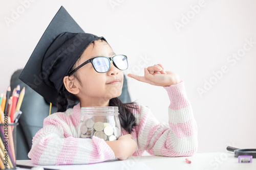 Little Asian girl wearing graduate hat putting the coin into clear glass jar piggy bank and smile with happiness for money saving to wealthness succesful in the future of education concept photo