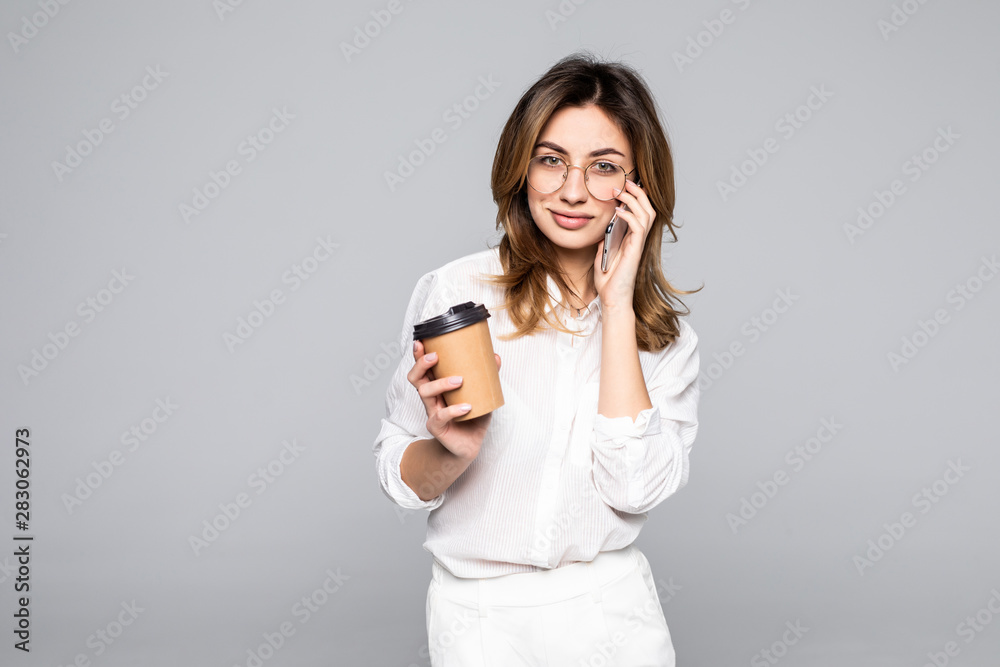 Young business woman posing talking by mobile phone drinking coffee isolated over grey wall background Young business woman posing talking by mobile phone drinking coffee isolated over grey wall background