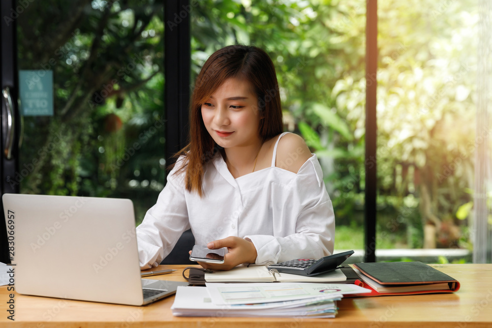 Young asian woman in smart casual wear holding smart phone and working on laptop while sitting near window in home office. Young asian woman in smart casual wear holding smart phone and working on laptop while sitting near window in home office.