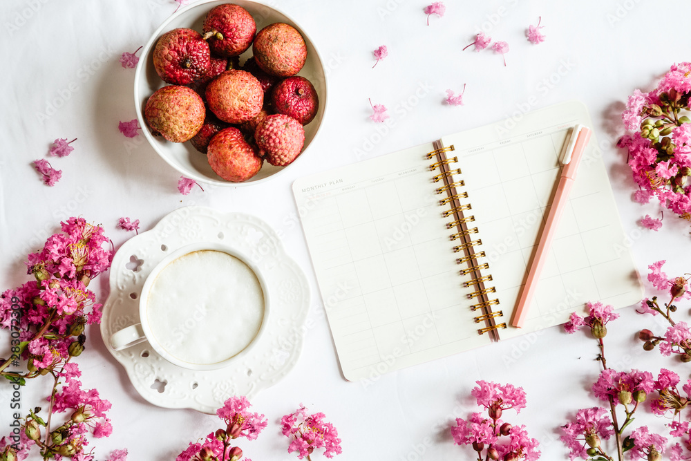 Morning coffee mug for breakfast, fresh lichee fruit, empty notebook, pen and pink flowers on white wooden table, top view, flat lay style. Woman working desk. Mockup, overhead