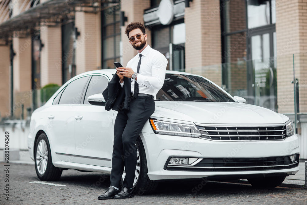 Handsome, smiling, happy, bearded manager is using smartphone and standing near his new white car on the street near the modern office center. Looking to the camera.