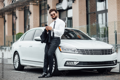 Handsome, smiling, happy, bearded manager is using smartphone and standing near his new white car on the street near the modern office center. Looking to the camera. © Тарас Нагирняк