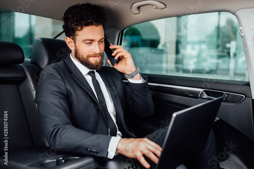 Handsome, bearded, smiling businessman working on his laptopand speaking mobile phone on the backseat of the car. © Тарас Нагирняк