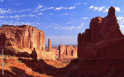 A giant rock formation dasts its shdow across a small valley in Arches National Park. Above, a blue sky is dotted with small puffy white clouds.