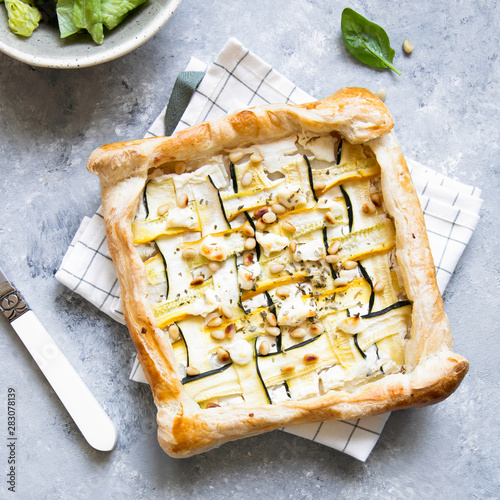 Courgette puff pastry pies with goat cheese and pine nuts on grey background.