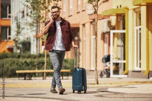 Ready for trip. Cheerful bearded man in casual wear and eyeglasses pulling his luggage and talking by phone while walking through the city street
