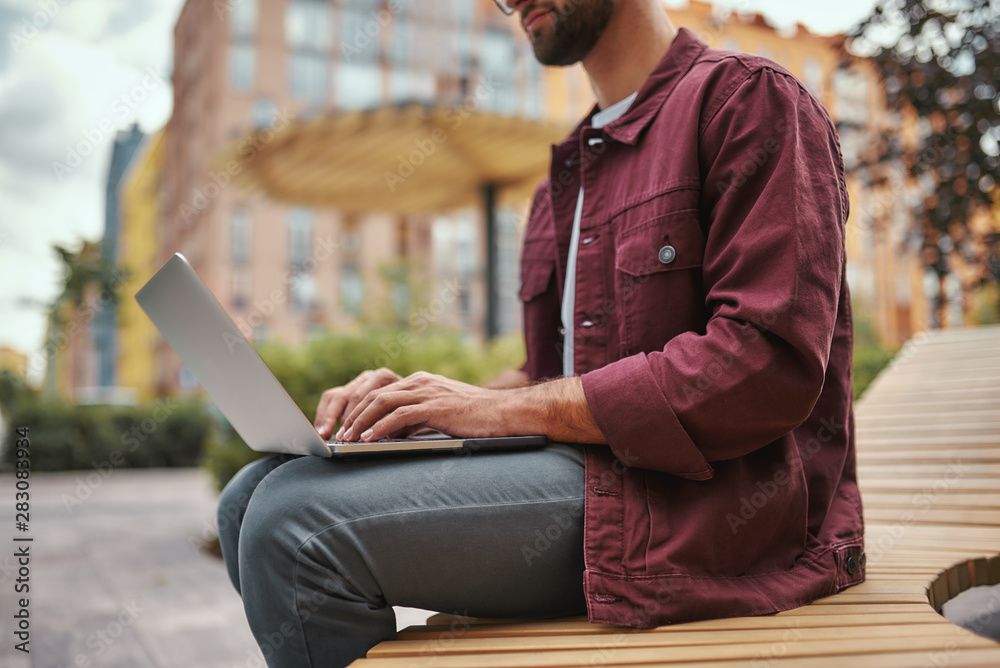 Free wi fi. Cropped photo of young handsome man with stubble in casual clothes working on laptop while sitting on the bench outdoors