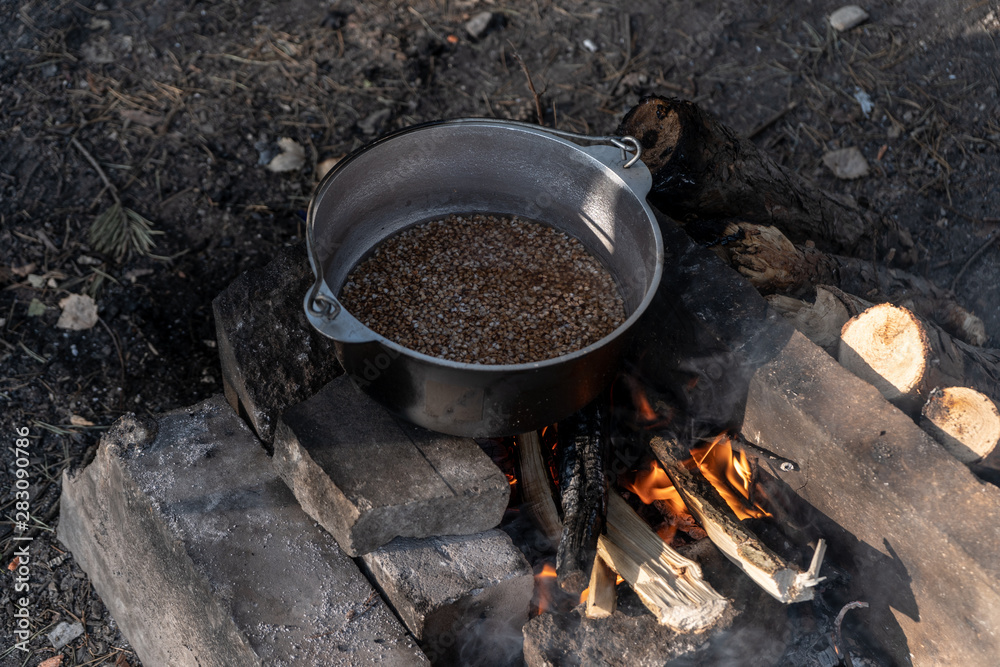 Buckwheat in a cauldron is prepared on a campfire on a sunny summer morning in the forest. Top-side view. Buckwheat in a cauldron is prepared on a campfire on a sunny summer morning in the forest. Top-side view.