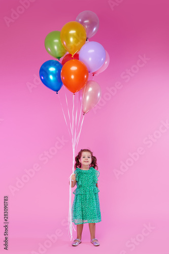 Portrait of a cheerful little toddler girl over pink background, holding bunch of colorful air balloons. The concept of children holiday birthday