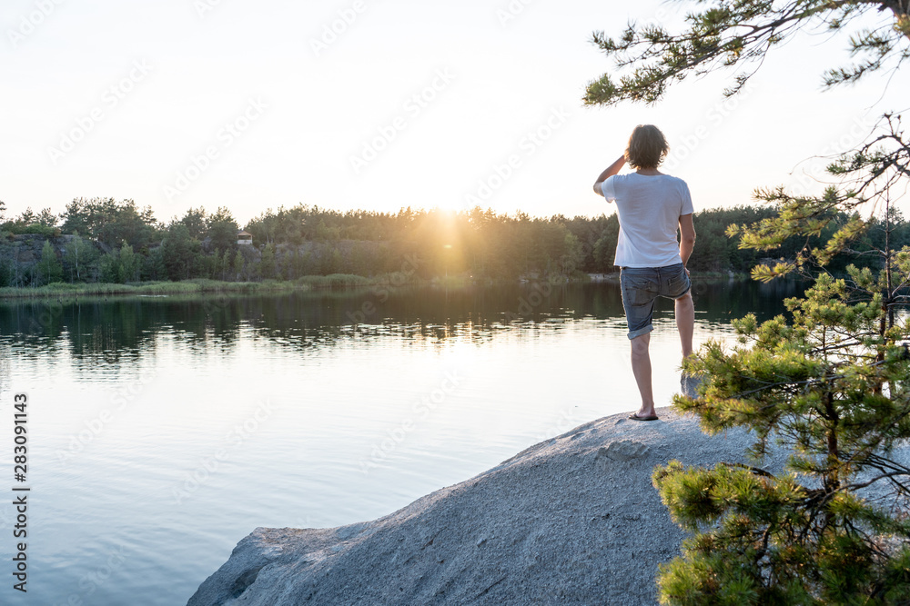 A young man stands on the cliff above a quarry filled with clear blue water, in the summer at sunset. Picturesque natural landscape