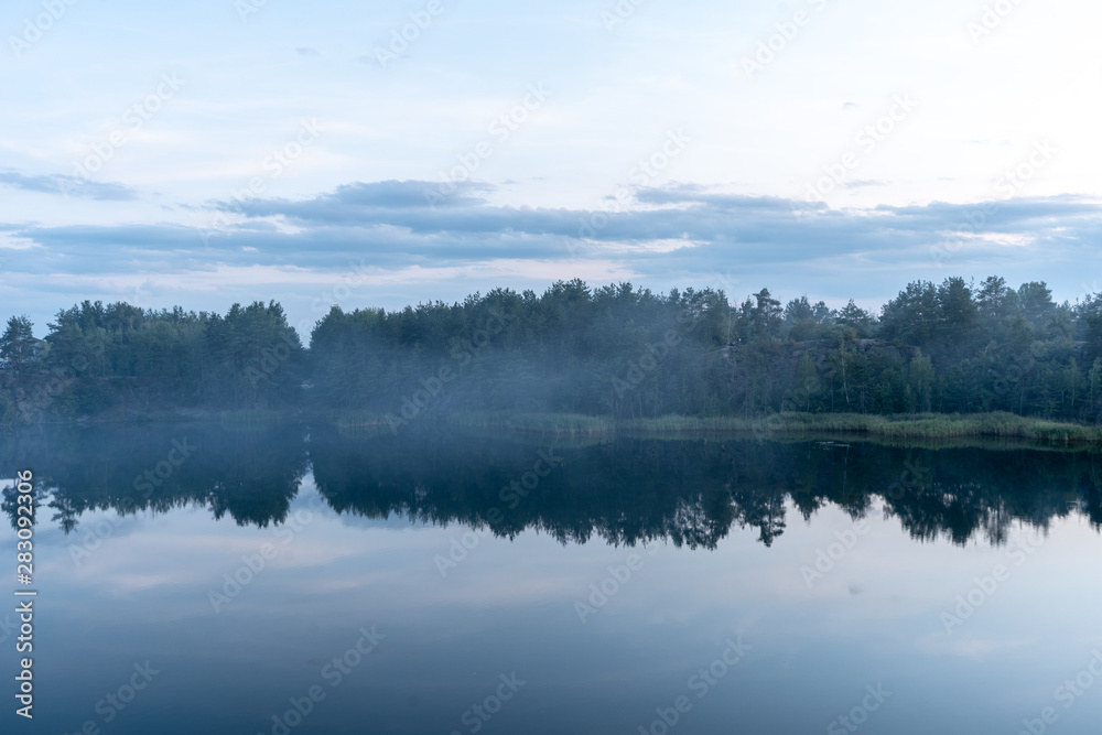 Dramatic scenic view of the granite quarry filled with blue water, and the opposite shore overgrown with trees after sunset at dusk in the summer. 