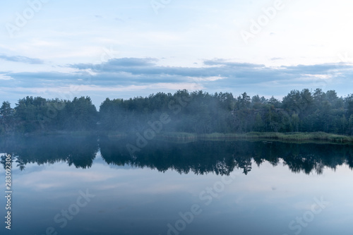 Dramatic scenic view of the granite quarry filled with blue water, and the opposite shore overgrown with trees after sunset at dusk in the summer.  © MoonfliesPhoto