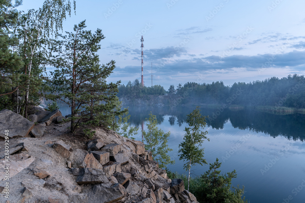 View of the granite quarry through stones filled with water, and the opposite rocky shore overgrown with trees after sunset at dusk in the summer. High tower on the opposite bank