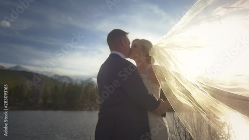 A bride’s veil blows across the frame as she embraces her beloved by a lake