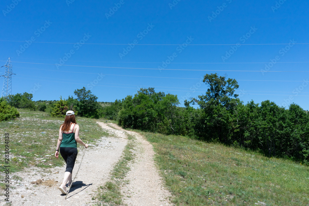 Girl hiker walking along a pathway with her backpack. Sport caucasian woman jogging in summer with hat and wood stick. Red head Fit vitality female making exercise in a journey
