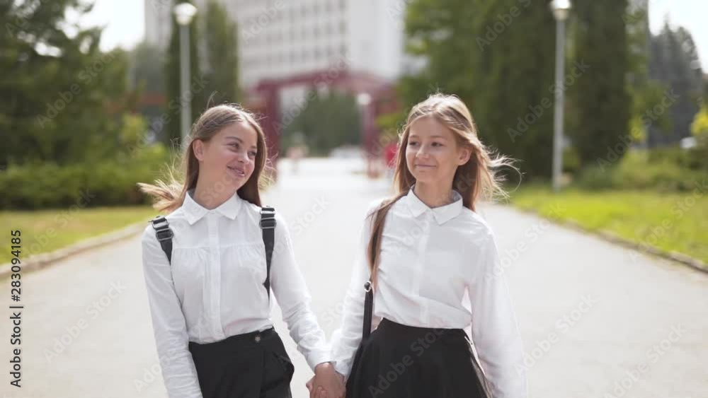 Two happy young girl students walk together towards frame on sunny day