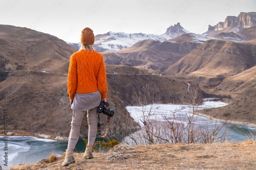 Portrait from the back of the girl traveler photographer in an orange sweater and hat with a camera in hand in the mountains against the background of a frozen mountain lake. Photo travel concept Portrait from the back of the girl traveler photographer in an orange sweater and hat with a camera in hand in the mountains against the background of a frozen mountain lake. Photo travel concept
