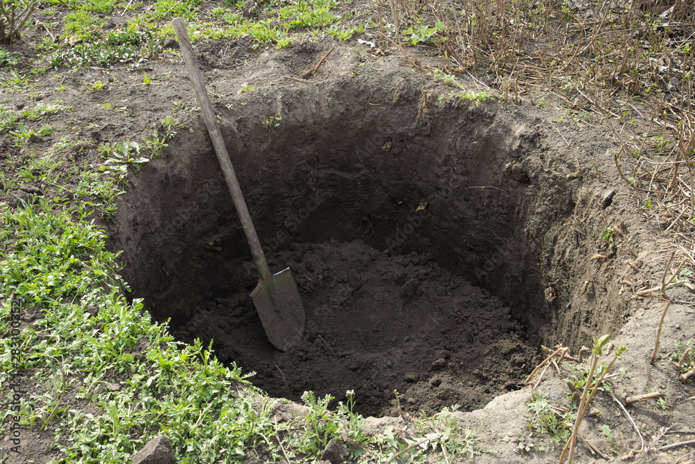 Deep pit in the ground. In the pit lies a shovel. Digging a hole. - obrazy, fototapety, plakaty 