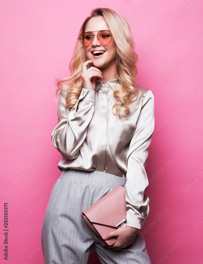 Young beautiful blond woman wearing blouse, pants and sunglasses holds a handbag and posing on a pink background. Young beautiful blond woman wearing blouse, pants and sunglasses holds a handbag and posing on a pink background.