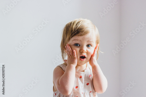 Leinwand Poster Surprised shocked child toddler girl with hands on her cheeks isolated on light