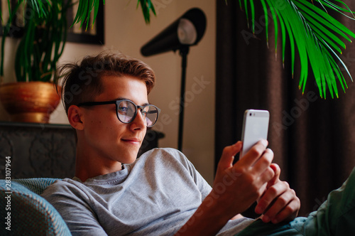 Smiling boy in glasses using smartphone sitting in the chair indoor photo