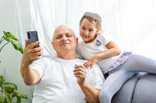 Portrait of the cute girl playing or typing on the smartphone while sitting on the couch with her grandpa at home.