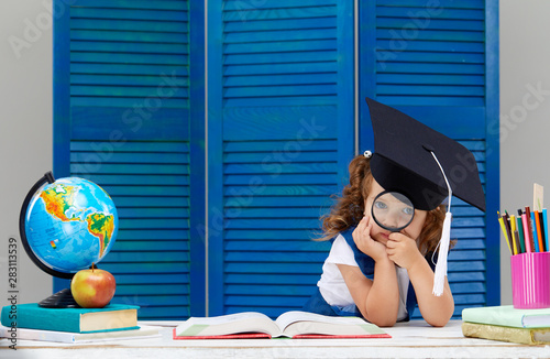 Clever and smart child, prodigy. A little girl in a graduation cap is examines a schoolbook through a magnifying glass. Sitting at a desk with books on blue wooden wall background