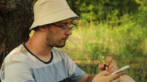 young handsome caucasian naturalist scientist sitting under a tree on the grass, smoking a cigarette and making notes in a notebook, medium plan photo