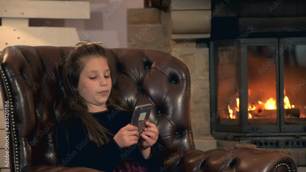 Little girl spend her leisure time with smartphone near the fireplace and sitting on a big comfortable armchair at home in the evening