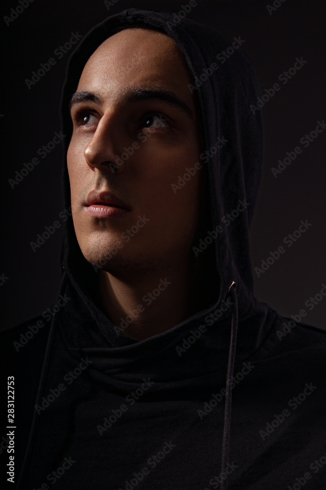 Mysterious serious man in black hoodie with hood on the head looking up on dark background. Dangerous criminal person Mysterious serious man in black hoodie with hood on the head looking up on dark background. Dangerous criminal person