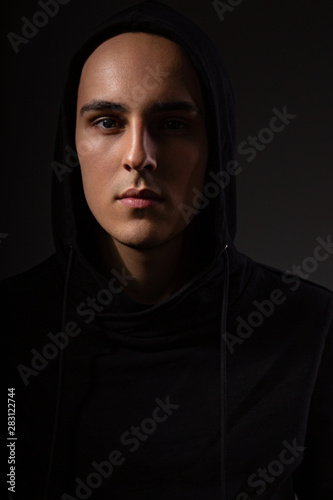 Mysterious angry man in black hoodie with hood on the head on dark background. Dangerous criminal person