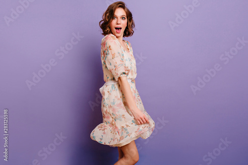 Studio shot of magnificent young lady with dark-brown hair plays with dress. Indoor portrait of surprised girl in attire with floral pattern dancing on purple background.