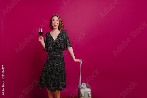 Lovely female model with glass of alcohol beverage posing in studio after travel. Stylish girl in elegant dress holding suitcase and drinking wine with pleasure. photo