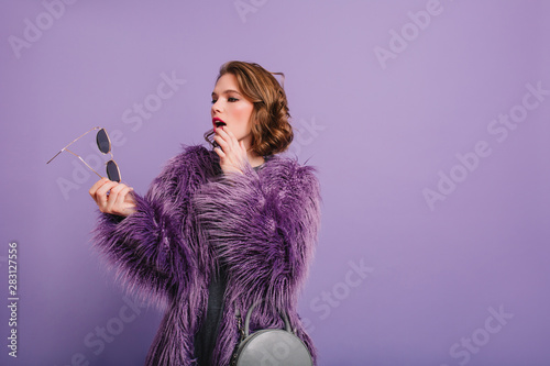 Serious woman in trendy purple jacket looking at her dark sunglasses in hand. Studio shot of pensive european girl in fur coat posing on indoor photoshoot.