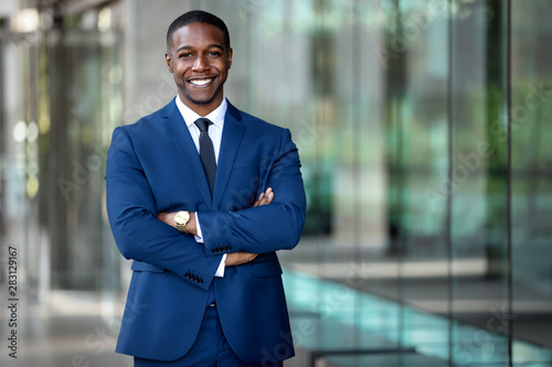 Smiling african american businessman CEO standing proud with arms crossed outside office workplace, colorful, reflective glass building, copy space