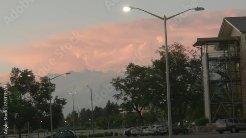 Timelapse of a distant thunderstorm rolling past a parking lot as the sun sets over it and with people walking in the distant background