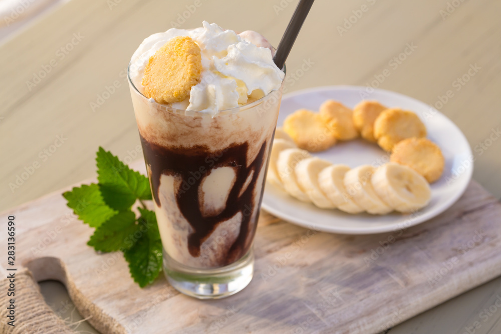 Tasty milkshake drink in glass mug with plastic tube and slice of banana on chopping board on wood table Tasty milkshake drink in glass mug with plastic tube and slice of banana on chopping board on wood table