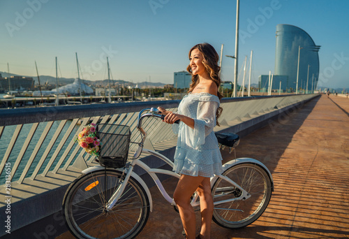 Beautiful Asian model in a blue dress rides a white bicycle in a modern city sea terrace yacht club on background. Concert of travel, ecology and health